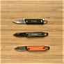 Three knives displayed open on a wood background. Orange handle in front; charcoal handle center; black handle on top.