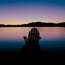 Barry Lobdell's photo card image of Adirondack chair facing water with Adirondack mountains in the distance. Sunset with purple and blue hues.