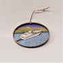 A white motorboat is centered in this oval ornament. It sits on a blue water background with a golden mountain and purple sky in the background.