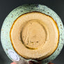 Unglazed circle bottom of mug with artist's imprint in the middle of a light-green colored speckled glaze
