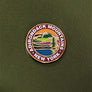 Close up of ADIRONDACK MOUNTAINS circular pin with colorful center circle depicting mountain, water and greenery. Outer circle has white type ADIRONDACK MOUNTAINS * NEW YORK. Pin is centered on the green card it is attached to.