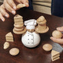 Fingers carefully holding tiny wooden loaf of bread aiming for the bakers pile on the left. The white-wooden rounded baker is centered on a wooden table with various tiny wooden baked goods surrounding him and another set of fingers holding a tiny wooden pie coming from the right.