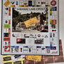 Game board of SARANAC LAKE-OPOLY open with game money on the board and below along with Merc Cards and Fate Cards featured below the board.