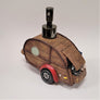 Woody camper soap dispenser slightly angled on a white background. The metal push spout sits atop the brown wood-grained look o fthe camper complete with hatch, circular window and red shield with wheel beneath.