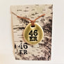 Ceramic, oval pendant with 46er in bold black in center and a taupe glaze around it on top of white. The ceramic pendant sits on a faux birch paper package with the brown necklace twine seen coming out of the birch card.