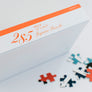 Side and bottom of puzzle box with orange lettering: 285 Piece 12" x 16.5" Jigsaw Puzzle with 6 loose pieces in front.