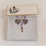 Small rectangular card with butterfly and flowers holds a pair of silver heart-shaped earrings that have MOM lettering positioned diagonally in the center hanging under a pale violet crystal bead. In the center between the earrings is a silver chain with a pale violet crystal bead hanging above the vertical lettering MOM. In all the MOM lettering the O is shaped like a heart. All sitting on white cotton in a white box with a white background.