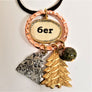 A copper circle independently surrounds a gold and silver outlined oval with 6er black type on white background. Attached to the copper circle are 3 charms: a sun coming out of a mountain, all silver metallic; a gold fir tree; a green-speckled stone ball with a gold circle on the bottom. A small part of the black necklace cord appears at the top.