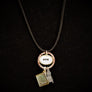 Hanging from partially seen black cord are a copper-colored necklace ring surrounding a gold oval framing a white oval with the black letters ADK printed centered in the oval. Hanging from the copper ring are three charms: visible here are the gold-framed map of the greater Saranac Lake area, a silver Adirondack chair. with a hint of the top of the third charm showing through in between the map and chair
