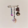 Allurement hatpin by itself on a white background. Hanging from pin is a multi-colored glass lure in blues, pinks, and greens; a pink feathered lure and a plain metal spinner.