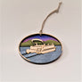 Natural wooden pontoon boat with four heads popping out is raised in the center of this oval ornament. Set on silver blue water with a green mountain and purple sky background.