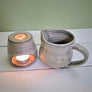 Ceramic maple syrup warmer pictured separated with top and bottom next to each other on a white planked surface with a green background. The bottom of the ceramic warmer is on the left with the lit tea light showing through the oval opening on the bottom and light showing through the small circular opening on the top. The mini ceramic pitcher sits to the right of the bottom.