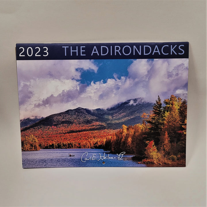 Front cover of 2023 Carl E. Heilman wall calendar. Navy bar at top reads 2023 THE ADIRONDACKS. Below that is a full-color photograph of autumn foliage in the foreground of the mountains covered with white fluffy clouds with blue sky peeking through. At the bottom is blue water with a single, very small boater in the middle.