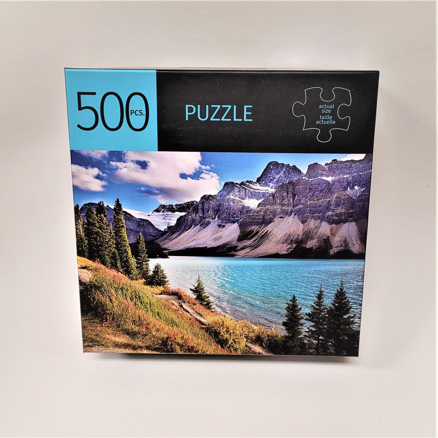 Cover of 500 PCS. puzzle with PUZZLE and actual size piece on top of the box cover and a full-color photo of snow-covered mountains, water and green landscape in the foreground of the puzzle photo.