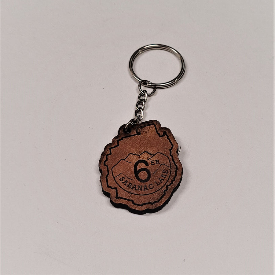 Single leather key chain with 6-ER SARANAC LAKE in brown lettering curved below a mountain range, all within the park outline, attached to a silver chain and keyring.