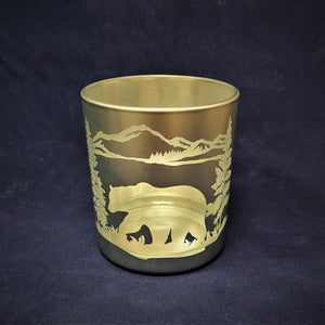 Glass candle holder on a blue background. Silver cup with etched out bear, evergreens, mountains and greenery.