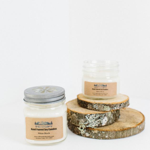 Left to right, jarred candle with silver lid, beige manufacturer's label with hint of white candle within. On 3 birch bark clabs sits an open candle jar. The manufacturer's label is displayed on the front of the open jar.