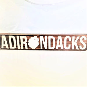 Brown horizontal sign with white lettering of the word Adirondacks. The O in the middle is in the shape of the Adirondack Park with a red heart in the upper portion.