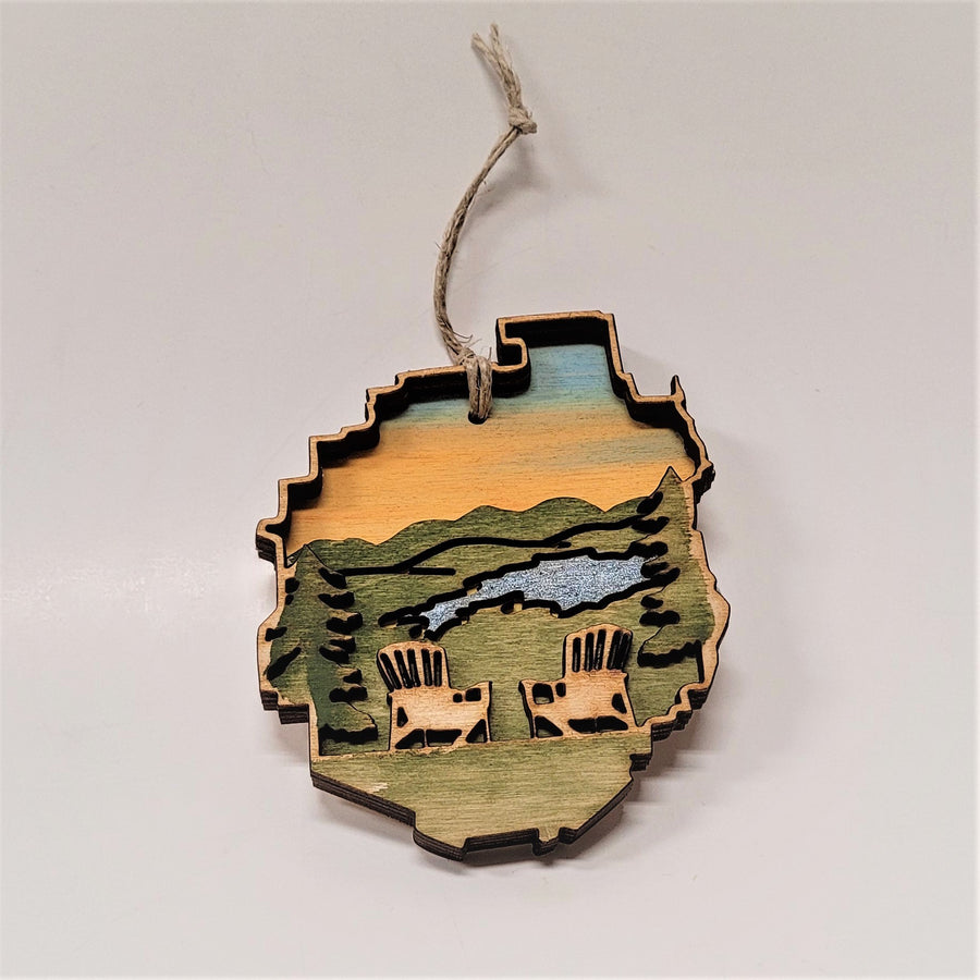 A pair of Adirondack chairs in the Adirondack Park ornament. The chairs are natural wood raised on a green background with a blue stream and golden sky. An evergreen is on the edge next to each chair.