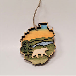 Bear ornament in the shape of the Adirondack Park. The bear is natural wood raised on a green background with a blue stream above, an evergreen to the left, green mountains and golden sky above.