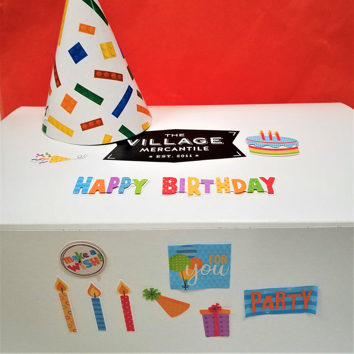 White box on an orange backdrop. Box is decorated with colorful birthday lettering and decals. A paper birthday cone hat sits on top of the box next to the Village Mercantile black and white logo decal.