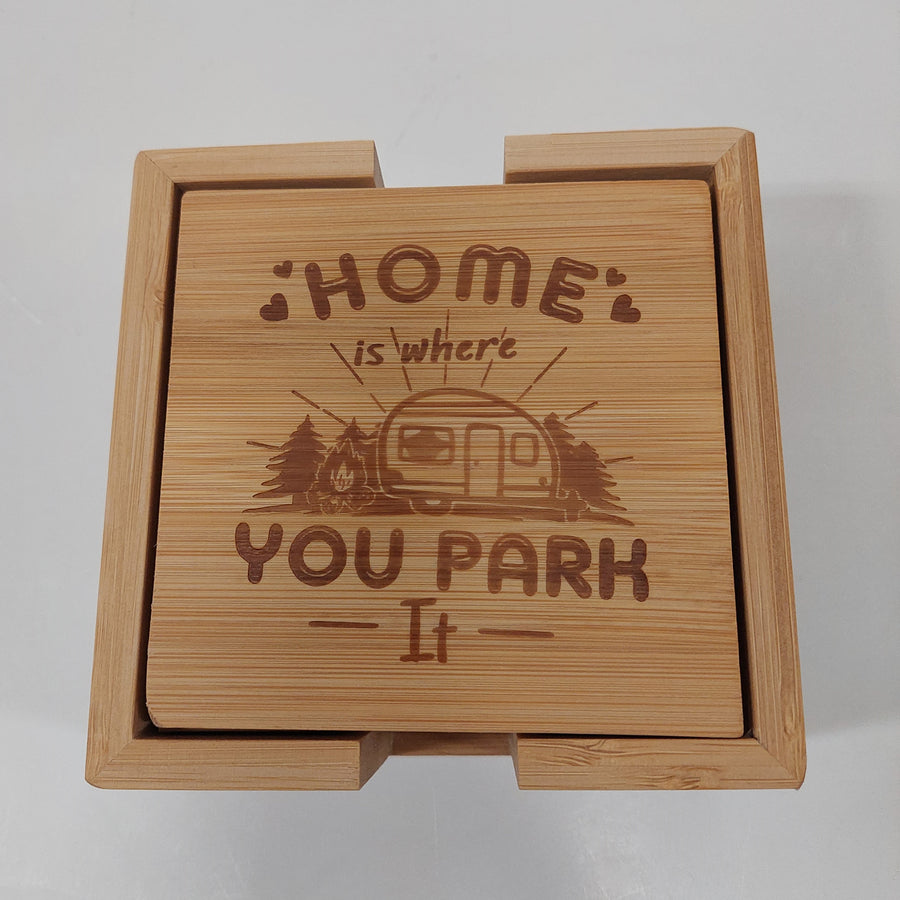 Square wood-like coaster with words Home is where You Park It etched above and below the image of a 50s camper with evergreens next to it. All in shades of brown.