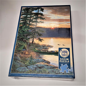 Cover of puzzle box featuring vertical photo of sunset on a lake with ducks in the water and a green canoe pulled up to a tent and campfire on a rocky shoreline. Type inset in blue box says Cobble Hill 500 PUZZLE * Poster Included