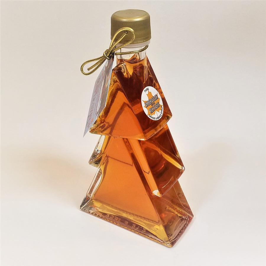 Clear glass Christmas Tree shaped maple syrup bottle filled with syrup. A gold-colored twist cap on top with a tag hung from a gold bow at the top left. A white circular label with a maple leaf sits on the uppermost right bough of the glass tree.