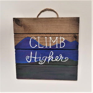 Wooden sign with twine hanging handle showing above. 4 wood panels with the bottom one painted green going in to the middle blue area with the top a natural wood. The words CLIMB Higher are painted in white in the center.