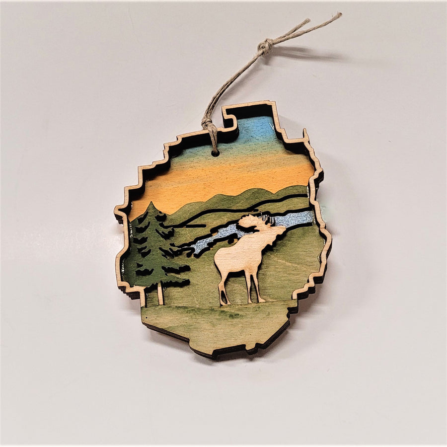 A natural wood raised moose is featured on this Adirondack Park ornament. An evergreen stands on the edge to the left. The background is green with a blue stream under a golden sky.