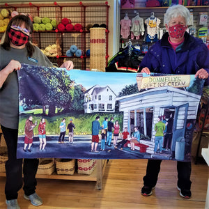 Inside the Village Mercantile, two women hold either end of the print of the iconic ice cream stand, Donnelly's, on a beach towel. White stand and house behind with people in solid colors ordering and eating.