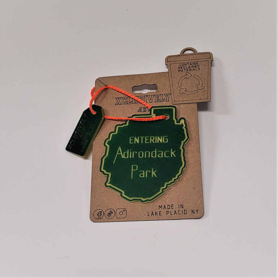 Single Adirondack Park ornament on its beige backing. An orange cord connects a Made in Lake Placid tag with the green ornament with yellow lettering ENTERING Adirondack Park.