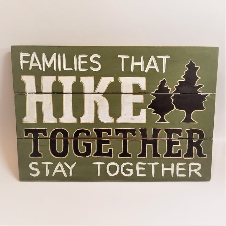 Green slatted sign. Top third has white test FAMILIES THAT, the top part of the word HIKE and the two of two black fir trees. Middle slat has bottom of the word HIKE in white, bottom of two black fir trees outlined in white and top of the word TOGETHER in black with white outline. The bottom third has the bottom of the TOGETHER lettering and white lettering STAY TOGETHER.
