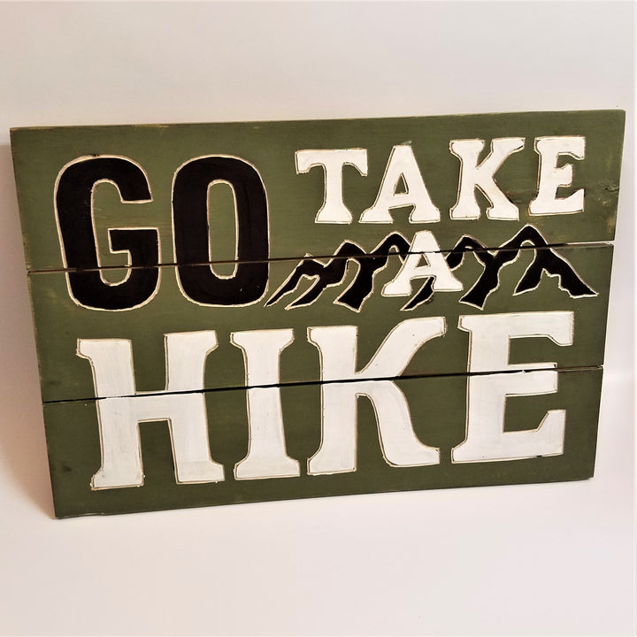 Three-paneled green sign. Top third has top of word GO in black type with white outline TAKE to its right with the top of Black peaks outlined in white and the top of the A; second panel has the bottom of GO, the bottom of the peaks and the A and the tope of the word HIKE in white type. Bottom third has the bottom of the word HIKE