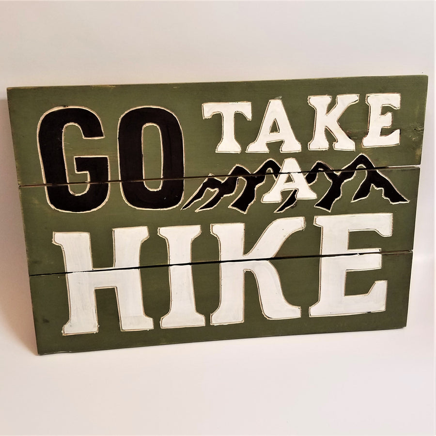 Three-paneled green sign. Top third has top of word GO in black type with white outline TAKE to its right with the top of Black peaks outlined in white and the top of the A; second panel has the bottom of GO, the bottom of the peaks and the A and the tope of the word HIKE in white type. Bottom third has the bottom of the word HIKE