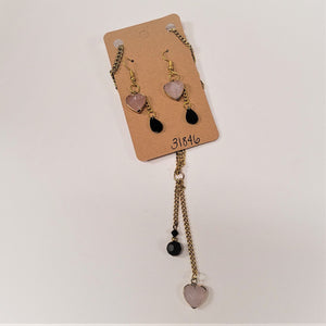 Heart-themed earrings and necklace hanging on and from a brown card. The earrings have a pale pink heart hanging from a gold hook above a black teardrop also hanging from a gold chain. Below the card the matching gold chain can be seen with a pale pink heart hanging from the bottom of a gold chain and above that a black faceted ball hanging from a gold chain. Both are attached to the gold ring above them.