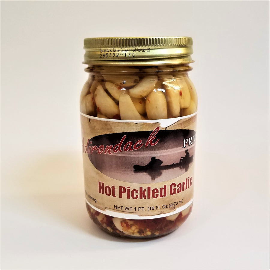 Glass jar of Hot Pickled Garlic with lots of peeled garlic cloves and specs of red and seeds showing through the jar around the label under the gold screw top.