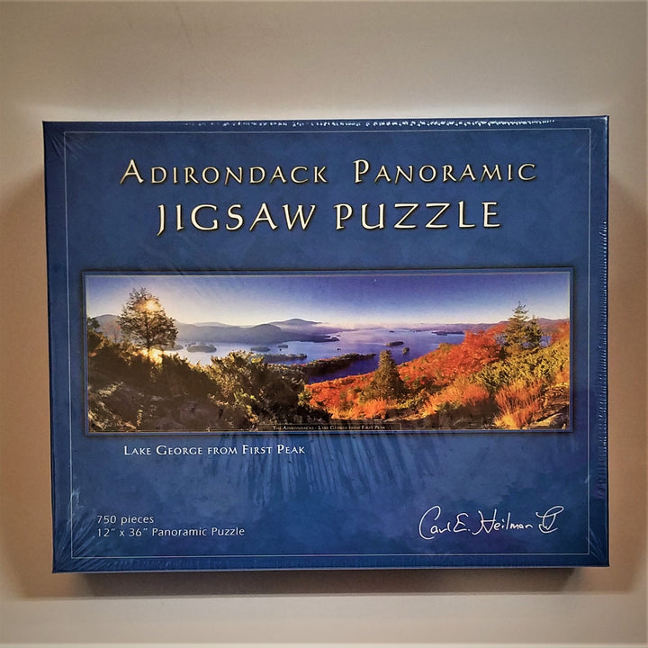 Cover of Adirondack Panoramic Jigsaw Puzzle: Lake George from First Peak. A blue border surrounds a horizontal photo of fall foliage facing the lake and purple mountains across the lake.