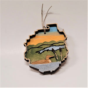 A brown loon is featured to the left in this Adirondack Park ornament with two cattails immediately to its right. All on a green background with a blue stream in the middle under a golden sky.