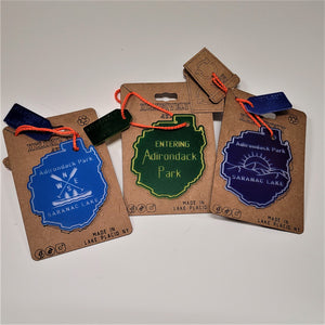Three Adirondack Park ornaments on their beige backing. Each has an orange cord with a Made in Lake Placid tag. Furthest left is sky blue with Adirondack Park above directional paddles , canoe, two fir trees and the lettering SARANAC LAKE; middle green ornament with yellow lettering ENTERING Adirondack Park; furthest right dark blue ornament with Adirondack Park Lettering above a sunrise over the lettering SARANAC LAKE