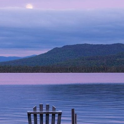 Image from Barry Lobdell's Meg's Moon photo card. Purple hues with an Adirondack chair facing blue and purple water and mountains under a white moon edging out of a purplish blue sky