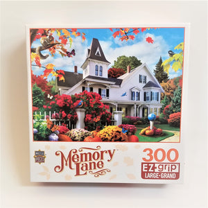 Box cover of Memory Lane 300 piece EZ grip jigsaw puzzle. Picture of a white two-story house with turret with full-color plants and manicured lawn surrounding it. Pumpkins, bluebird and butterflies also featured.