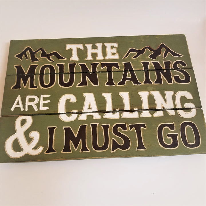 Green-painted 3-paneled wall sign. The text is in green and dark brown with THE on top next to mountain graphics above MOUNTAINS on one line ARE CALLING on the next line & I MUST GO on the last line.