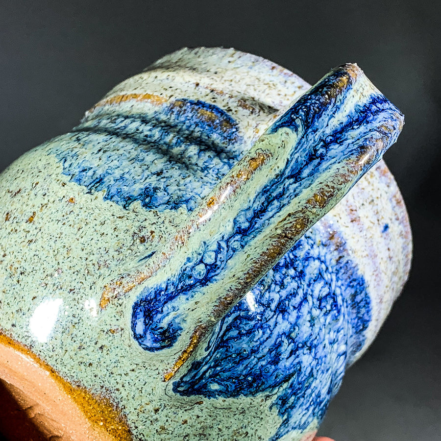 Diagonal view of ceramic mug with handle in middle. Blue speckled glaze on top left, handle and bottom right with green speckled glaze filling in elsewhere