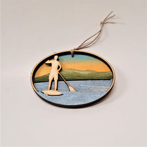 A natural wooden paddler standing is featured in the left side of this oval ornament. Below and around the paddler is silver blue water. In the background green mountains and golden sky.