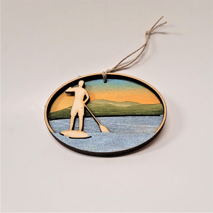 A natural wooden paddler standing is featured in the left side of this oval ornament. Below and around the paddler is silver blue water. In the background green mountains and golden sky.