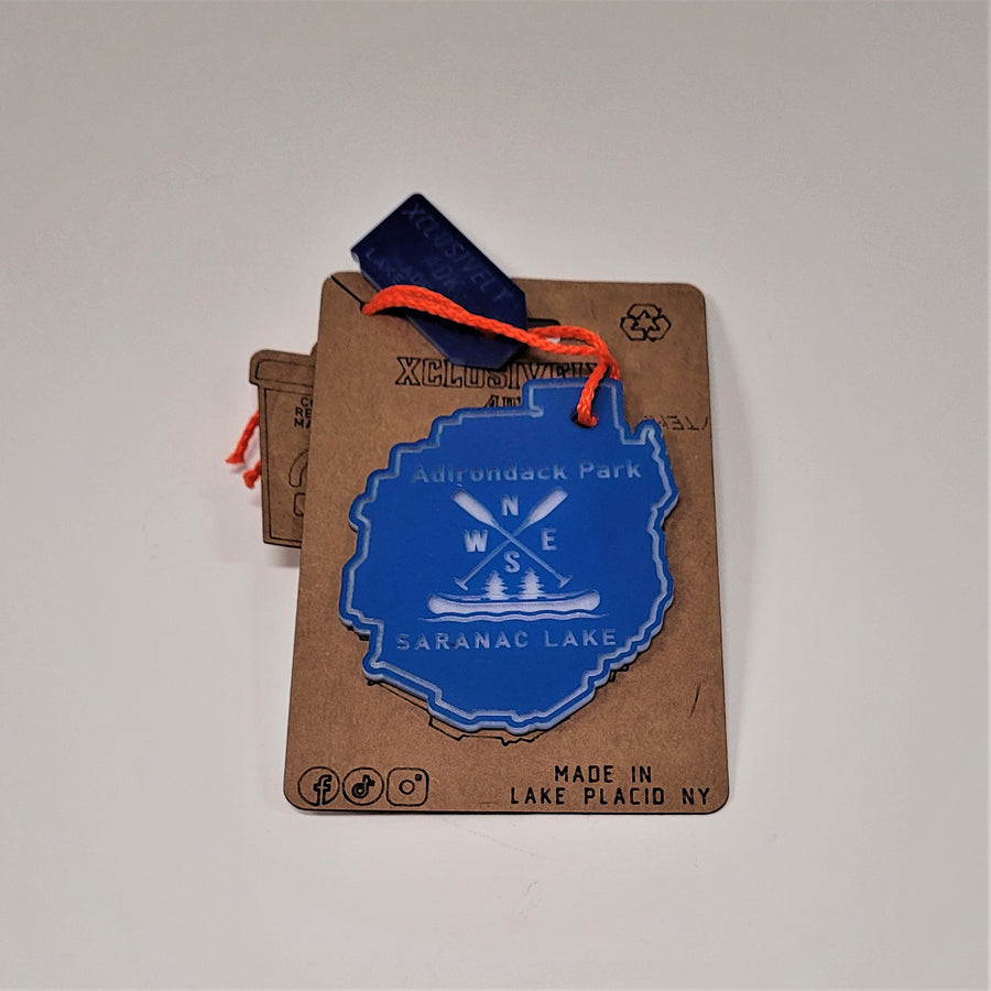 Single Adirondack Park shaped ornament on its beige backing. Sky blue with Adirondack Park lettering above directional paddles , canoe, two fir trees and the lettering SARANAC LAKE below.