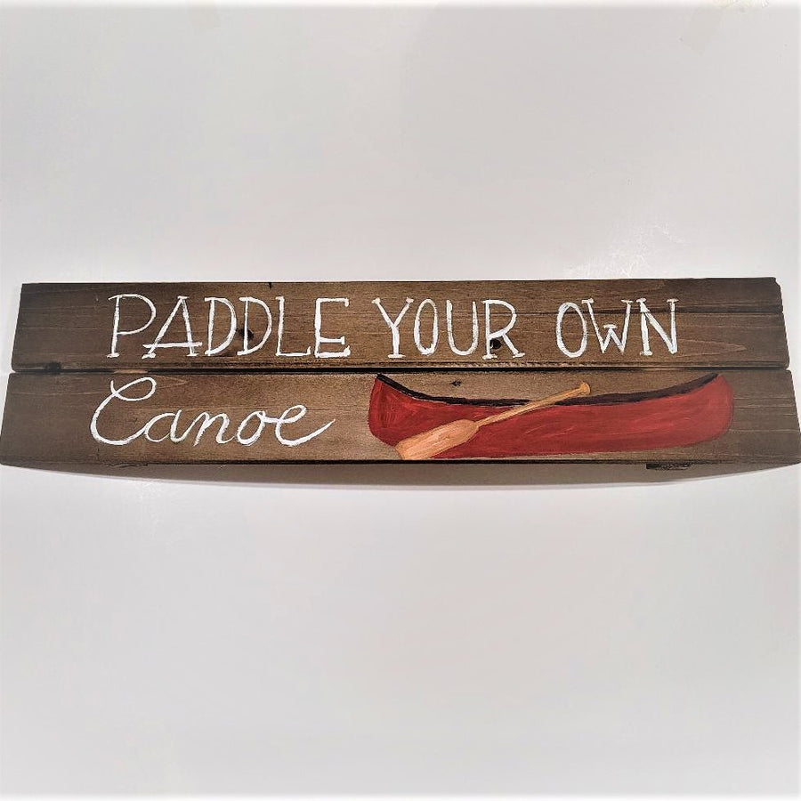 Wooden sign with two horizontal boards. On the top painted in white print are the words: PADDLE YOUR OWN. On the bottom panel the word Canoe is painted in script next to a red canoe with a light brown oar leaning on it.