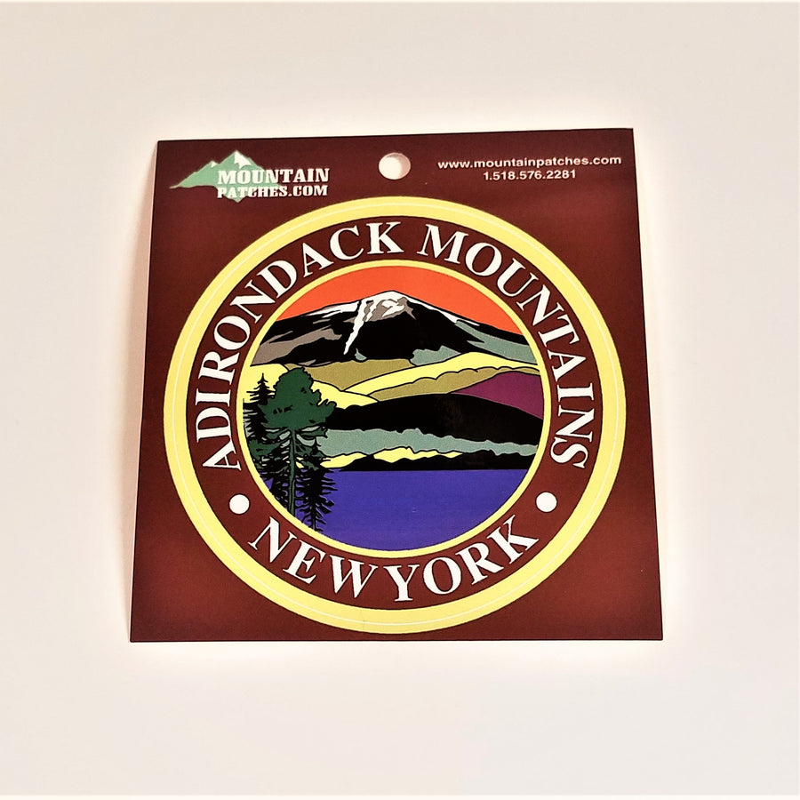 Full view of the Adirondack Mountains sticker on its brown packaging. A gold circle surrounds the white text: ADIRONDACK MOUNTAINS NEW YORK in the center circle an iconic color line art drawing of the mountains, water and evergreen.