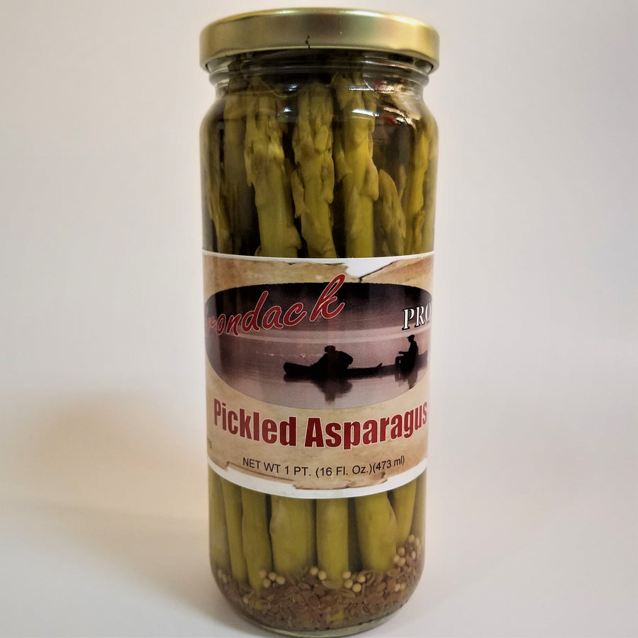 Glass jar of Pickled Asparagus with green stalks showing through the jar standing upright around the label under the gold screw top. Some seeds and garnish showing through the bottom under the label and the stalks.